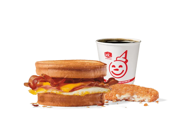 Image of the Supreme Sourdough Combo from Jack in the Box, showcasing a breakfast sandwich with crispy bacon, a fried egg, and melted cheese between toasted sourdough bread. Accompanied by a hash brown and a cup of coffee, all set against transparent background with the smiling Jack logo.