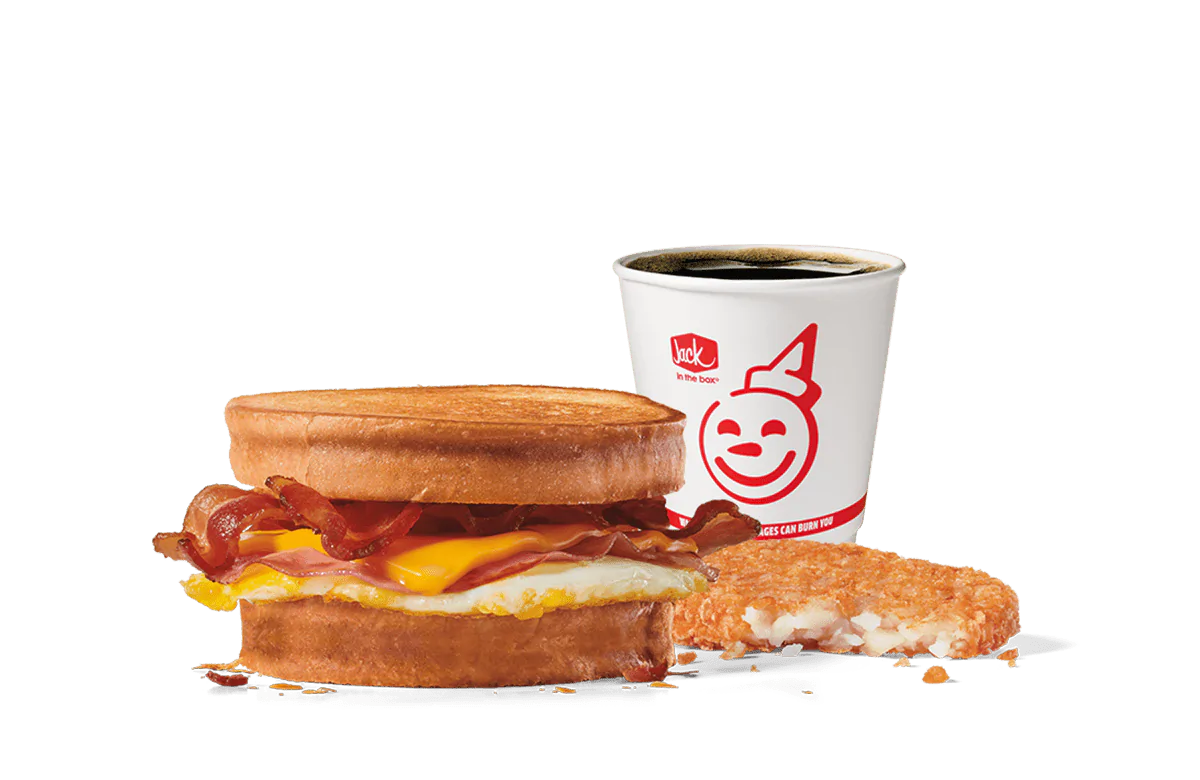 Image of the Supreme Sourdough Combo from Jack in the Box, showcasing a breakfast sandwich with crispy bacon, a fried egg, and melted cheese between toasted sourdough bread. Accompanied by a hash brown and a cup of coffee, all set against transparent background with the smiling Jack logo.